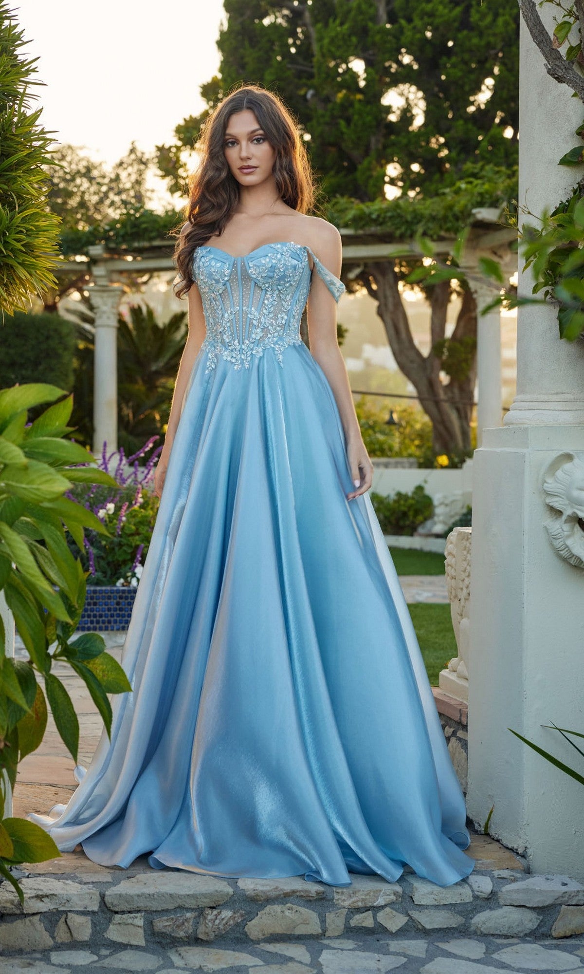 A woman stands outdoors in the Juliet JT2639G long prom dress, featuring a light blue off-shoulder corset bodice, lace details, and a flowing skirt amid lush greenery and elegant stone architecture.